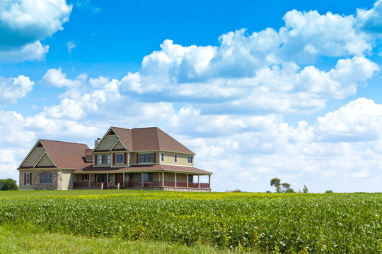 A large rural home with a wrap-around porch on a hill with a surrounding farm. 