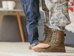 A child's bare feet standing on top of their parent's military boots. 