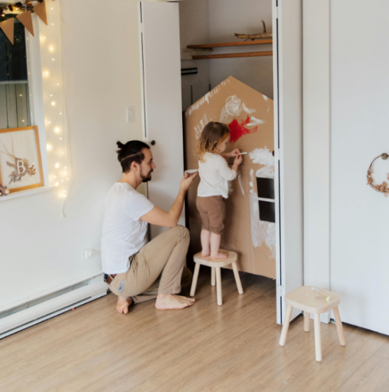 A father painting a dollhouse with his daughter standing on a stool.