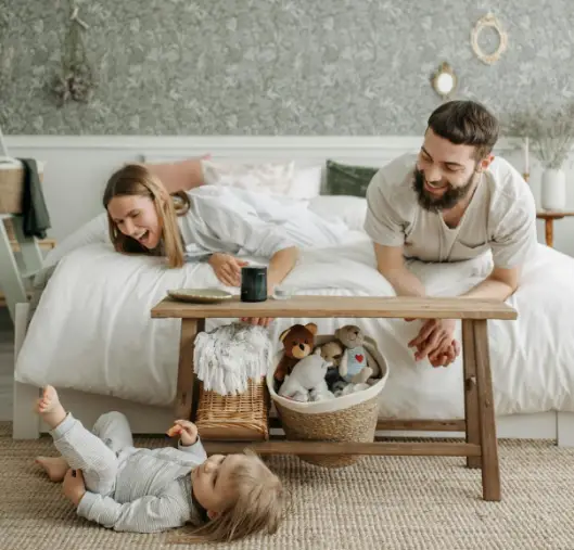 A mother and father at the foot of a bed looking at their toddler daughter on the floor, playing.