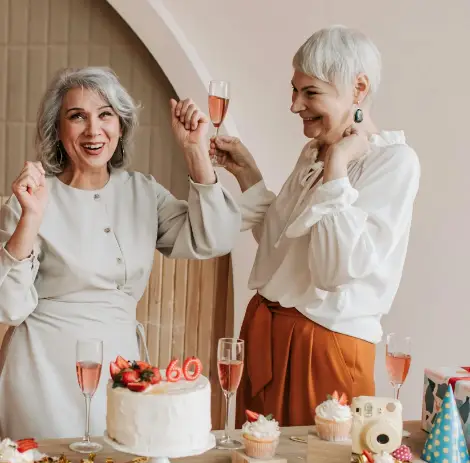 Two older women with gray hair celebrating with wine glasses and a white cake.
