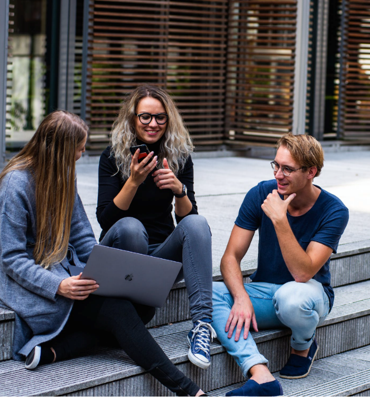 A group of friends (3) hanging out on the front steps of a building. One has their phone. Another has their laptop.