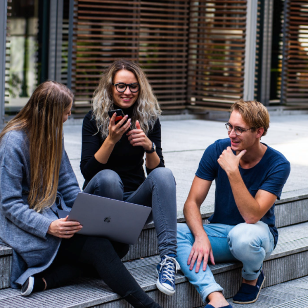A group of friends (3) hanging out on the front steps of a building. One has their phone. Another has their laptop.